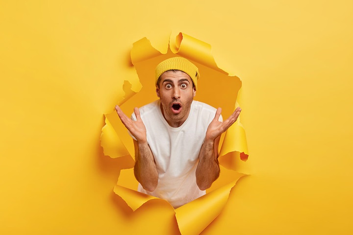 Surprised man spreads palms while breaking through a yellow wall of paper